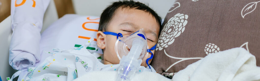 Young child lying in bed with an oxygen mask on, appearing to be asleep.